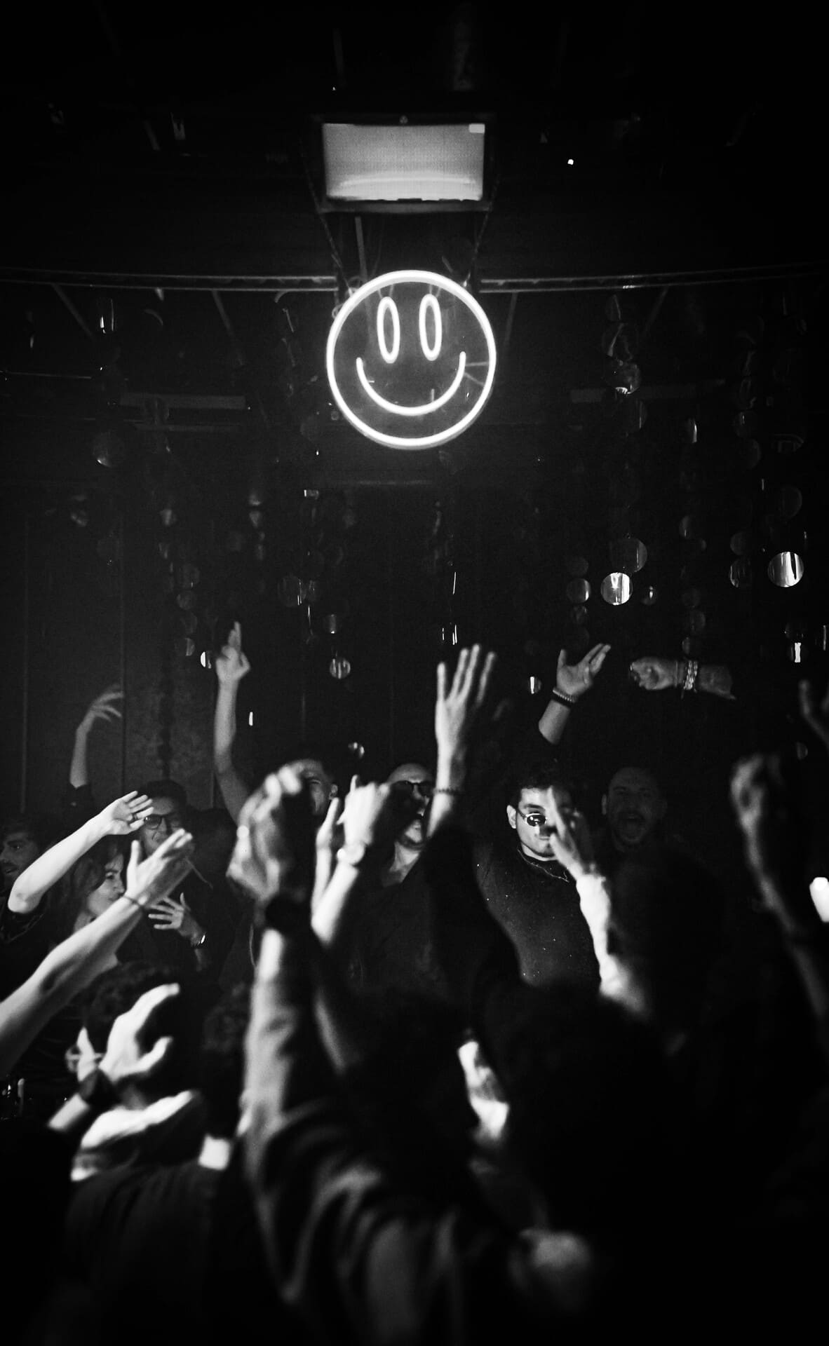Crowd dancing beneath a glowing smiley face neon sign in a dark nightclub.