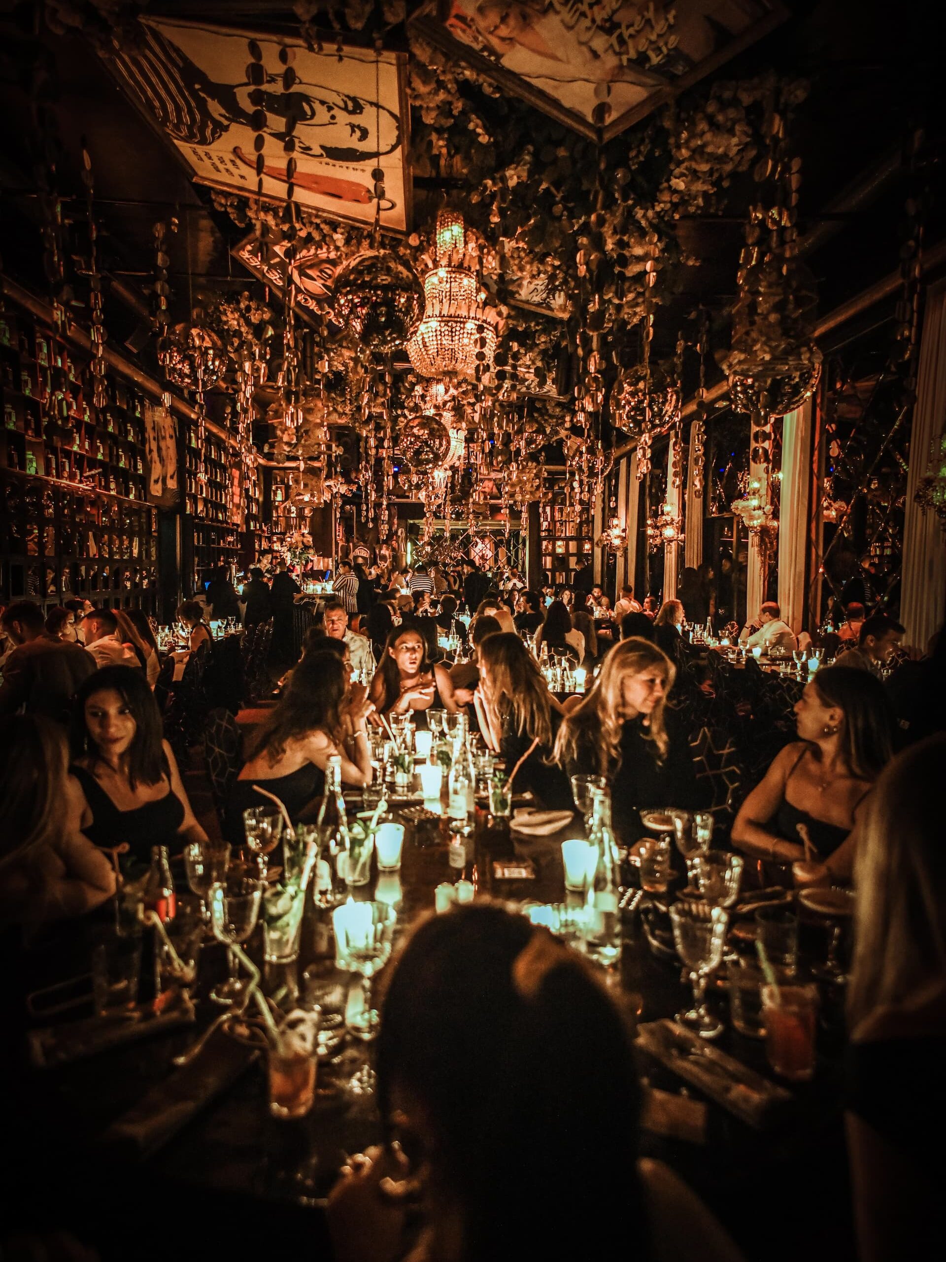 Guests dining at a candlelit restaurant with chandeliers and hanging decor during a lively evening