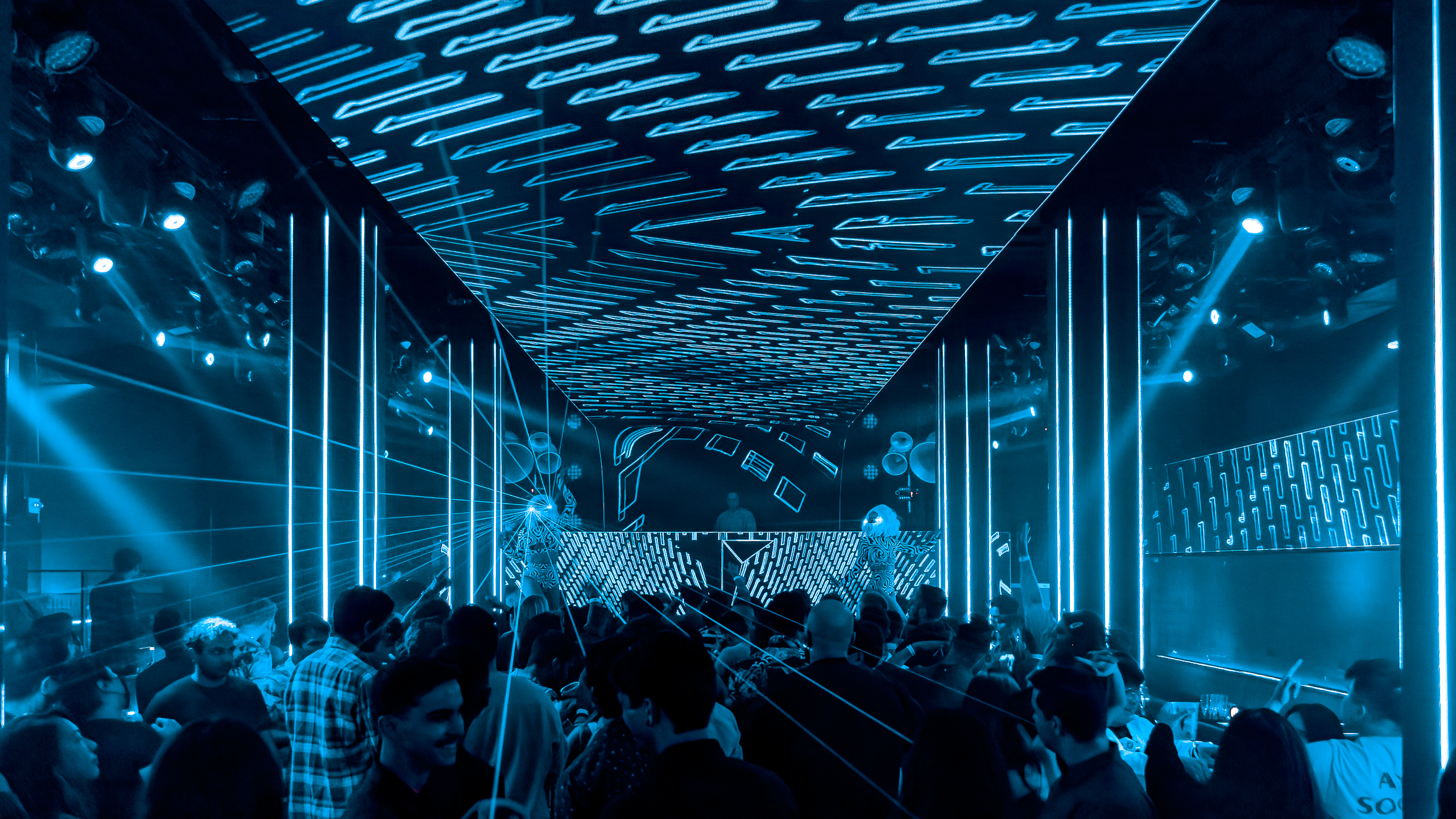 Crowd dancing in a modern nightclub with blue LED lights, geometric ceiling visuals, and a DJ performing on stage.