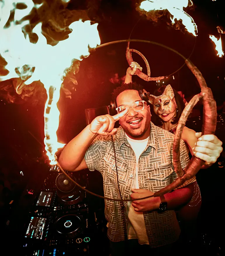 DJ posing behind turntables as a fire performer creates flames during a nightclub show.
