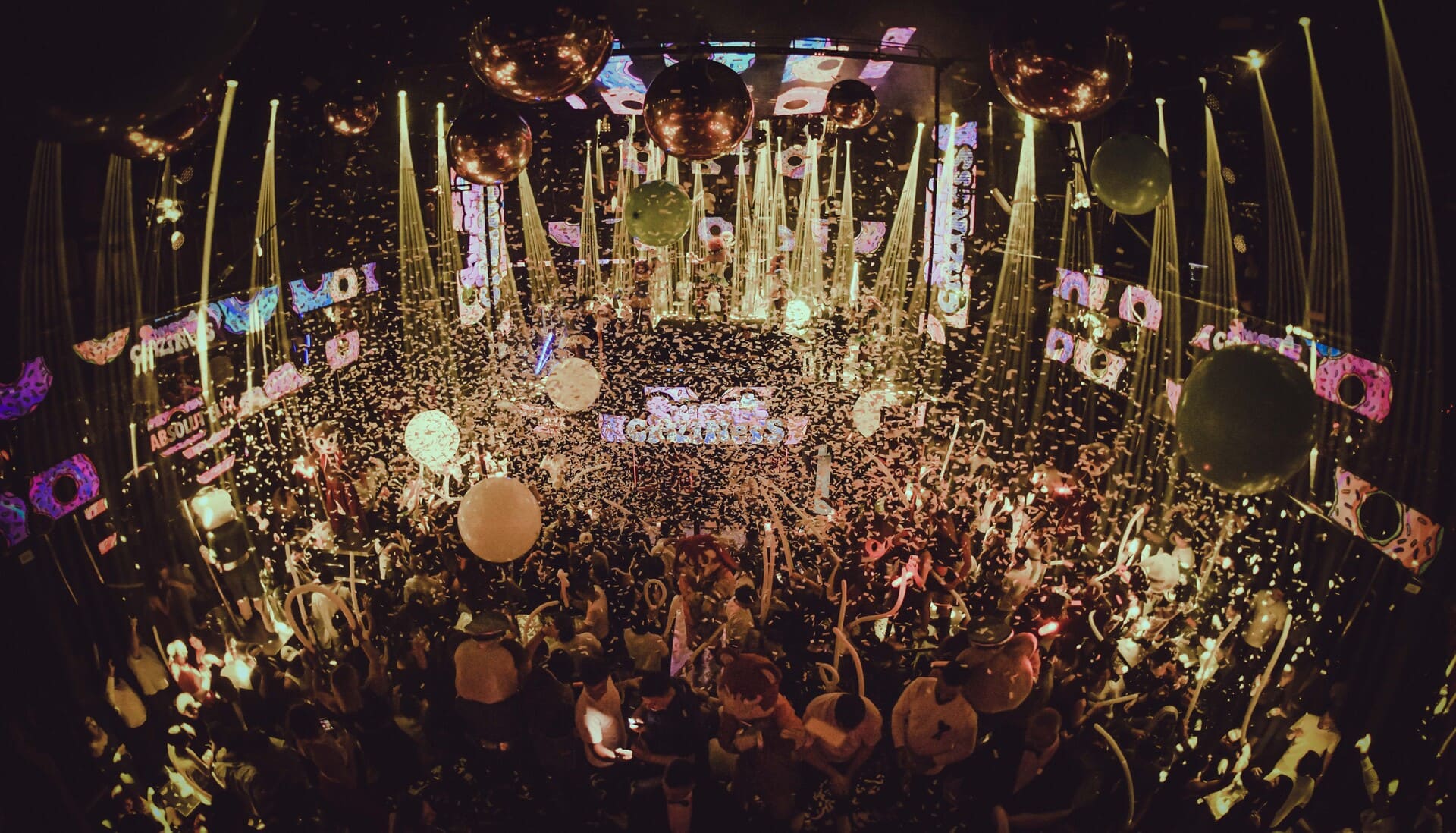 Wide aerial view of a nightclub packed with guests, balloons, disco balls, and falling confetti