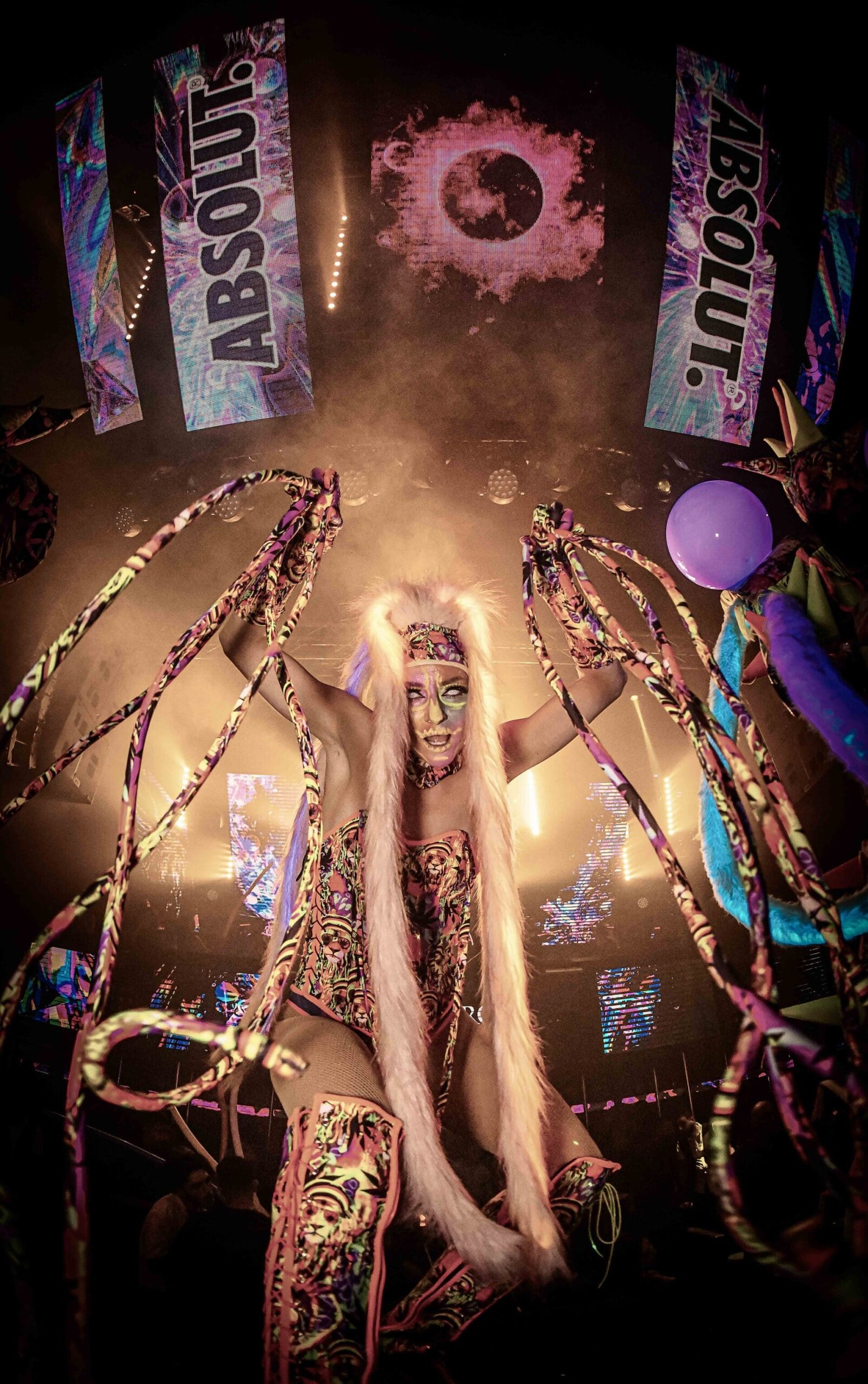 Performer in a colorful costume holding long ribbons beneath large LED screens in a nightclub
