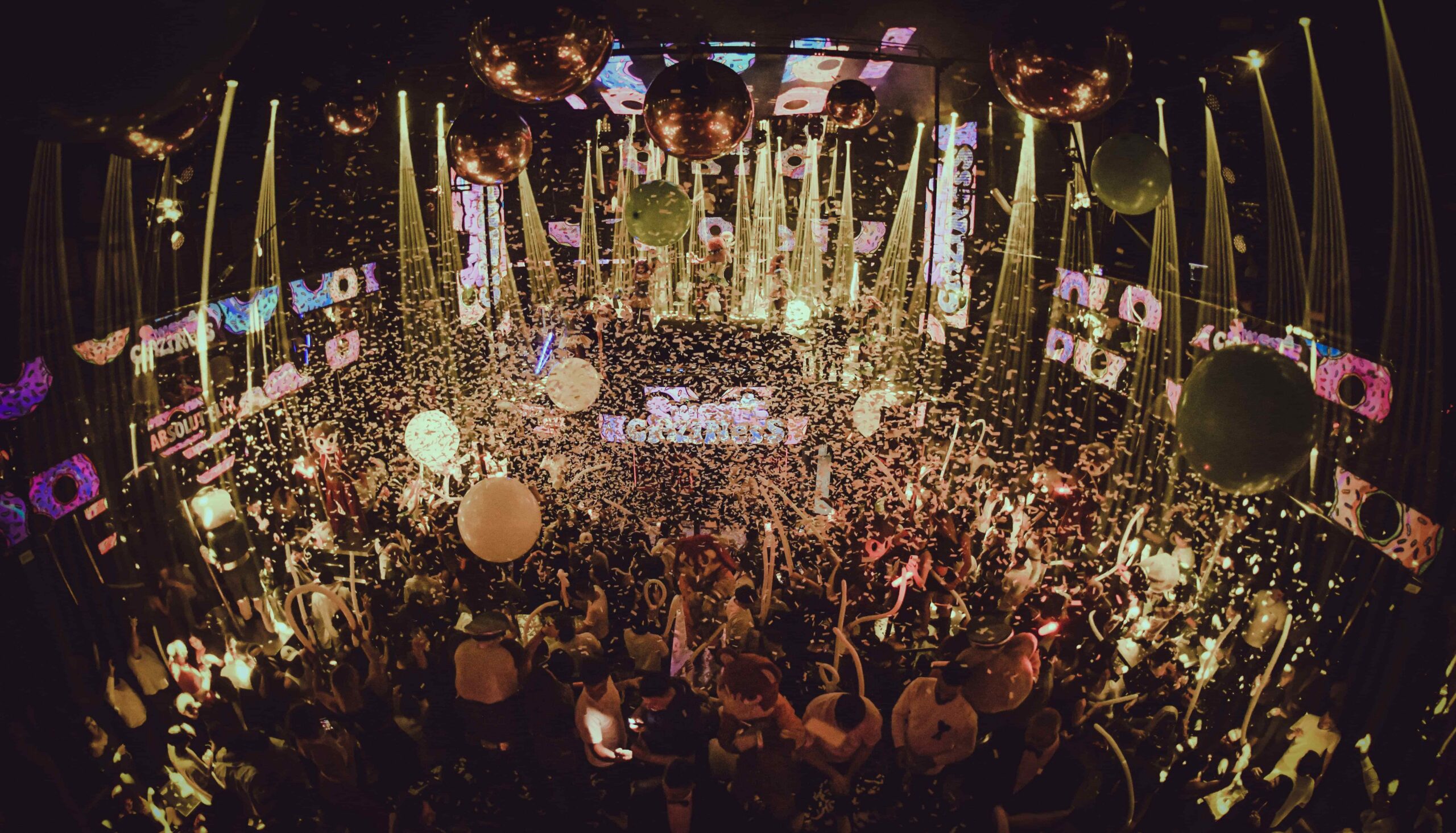 A crowded nightclub dance floor filled with people, falling confetti, large balloons, and golden streamers under bright stage lights and LED screens.